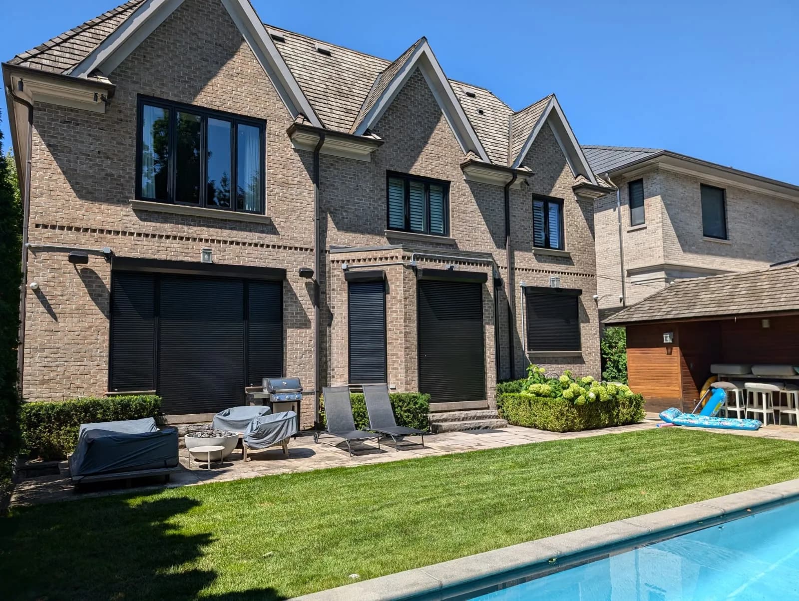 Modern Ontario brick home with multiple aluminum roll shutters deployed across the rear elevation, overlooking a pool