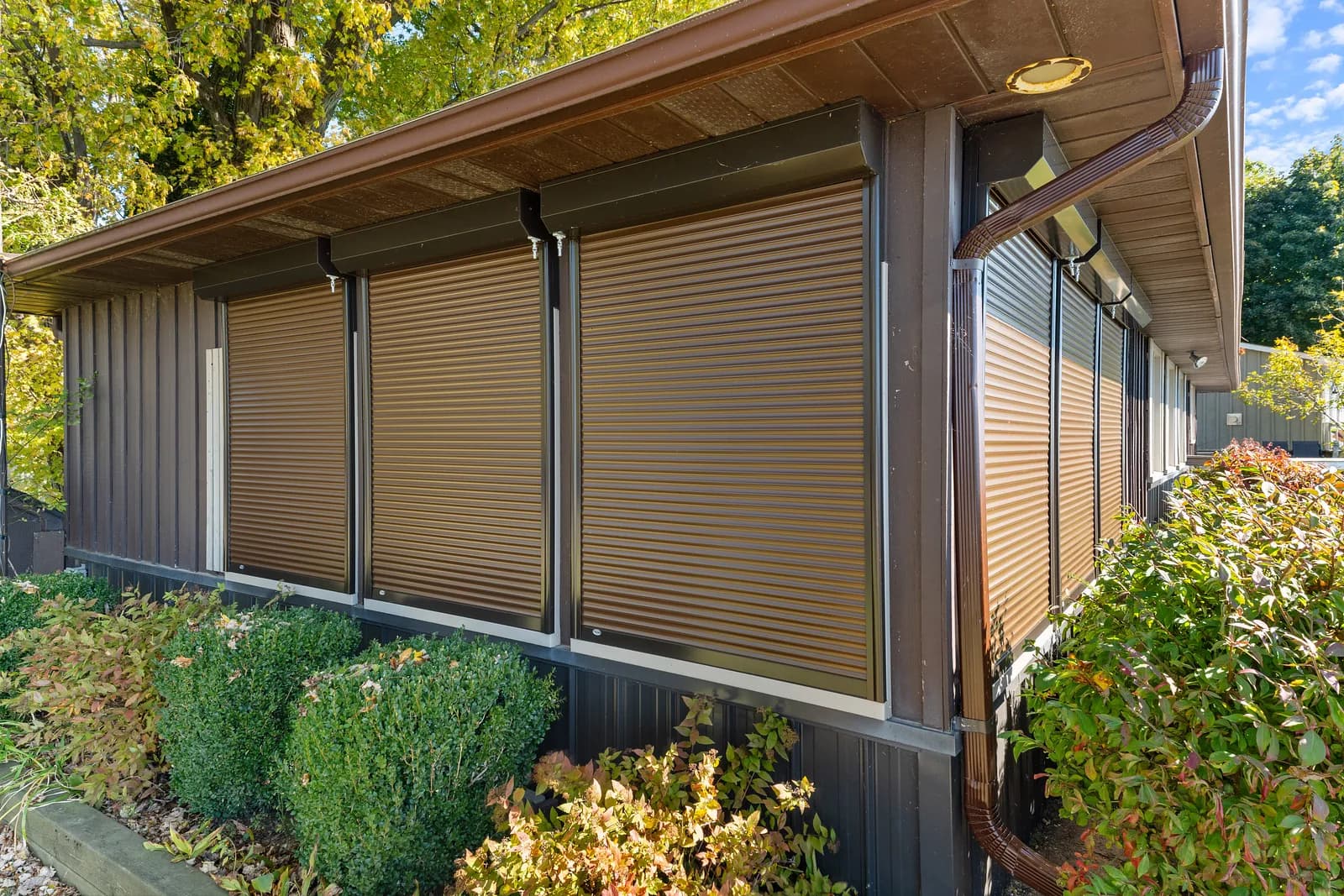 Three brown wood-grain aluminum roll shutters deployed across the side of a dark home
