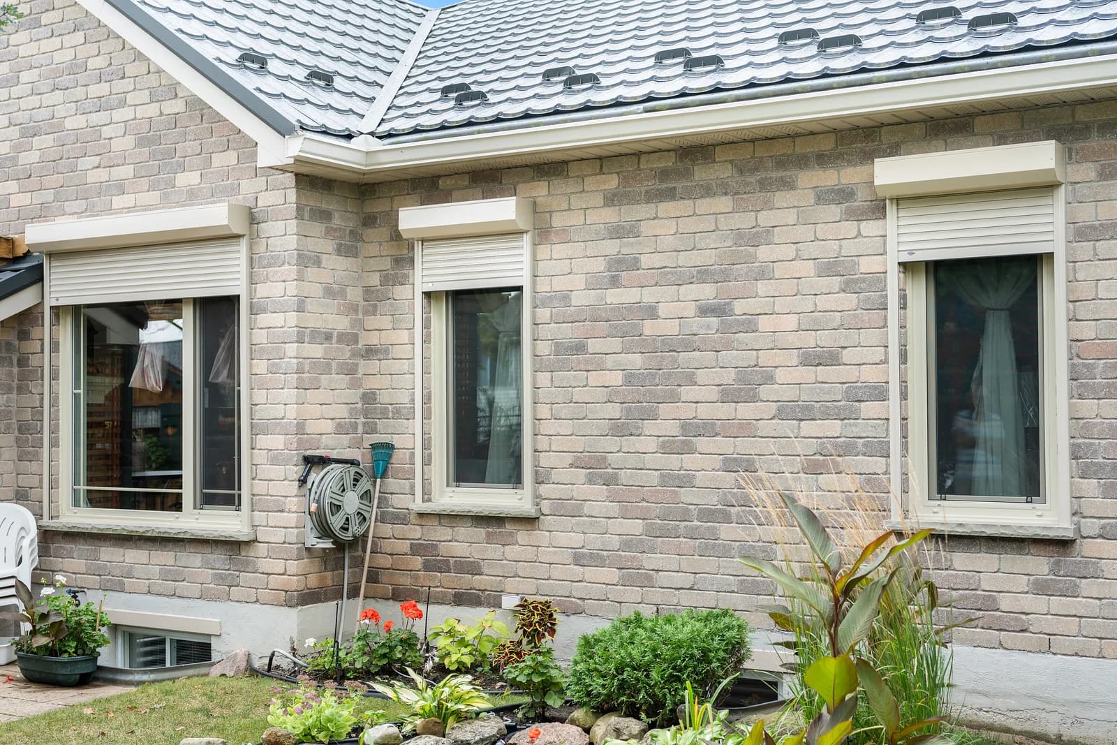 Light-brick bungalow with cream aluminum roll shutters retracted into headboxes above each window