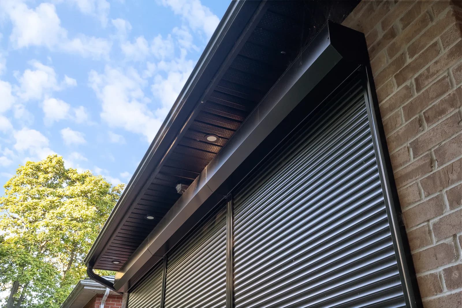 Black aluminum roll shutter deployed under a brown soffit with dramatic Ontario sky overhead