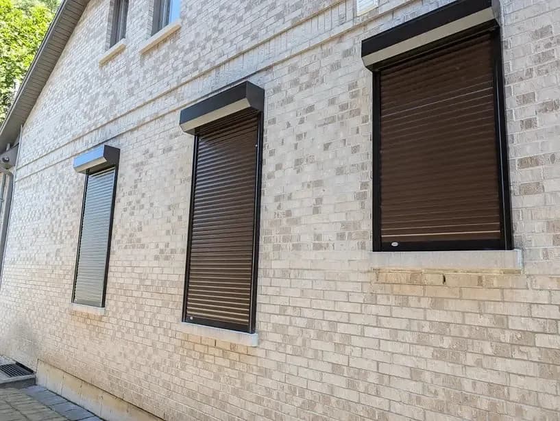 Two brown aluminum roll shutters deployed over windows on a light-brick exterior