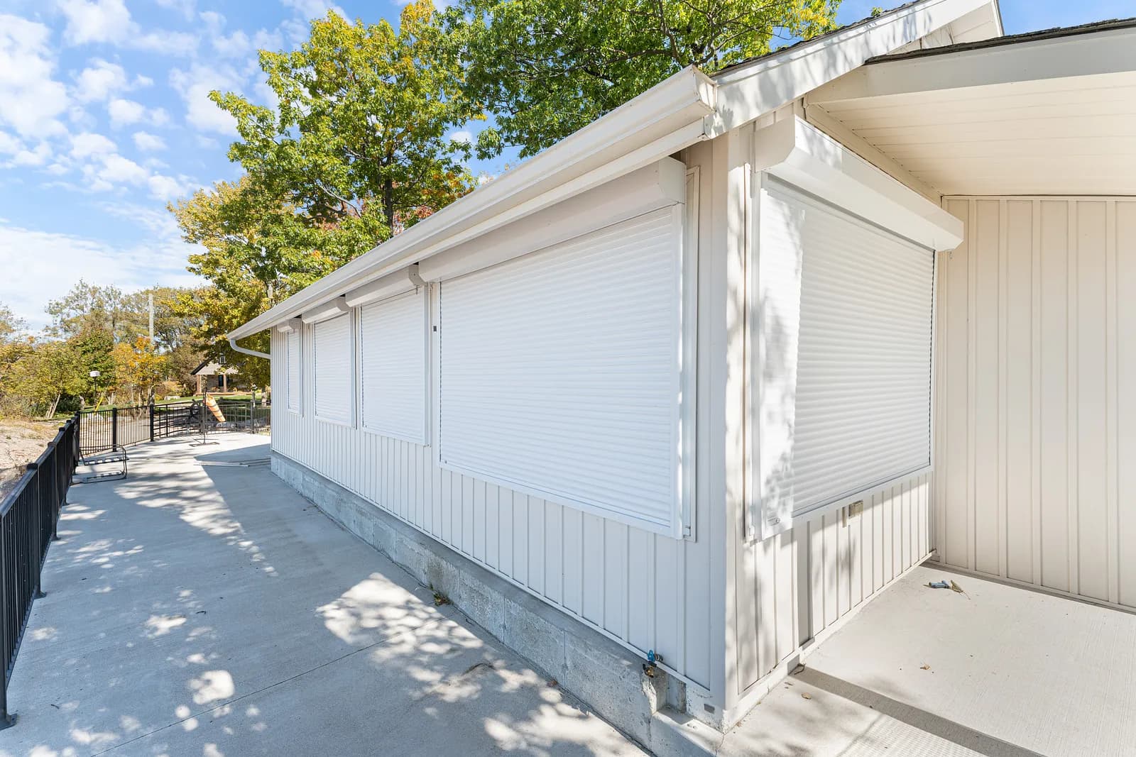 White-clad home side wall with white aluminum roll shutters deployed across a row of windows